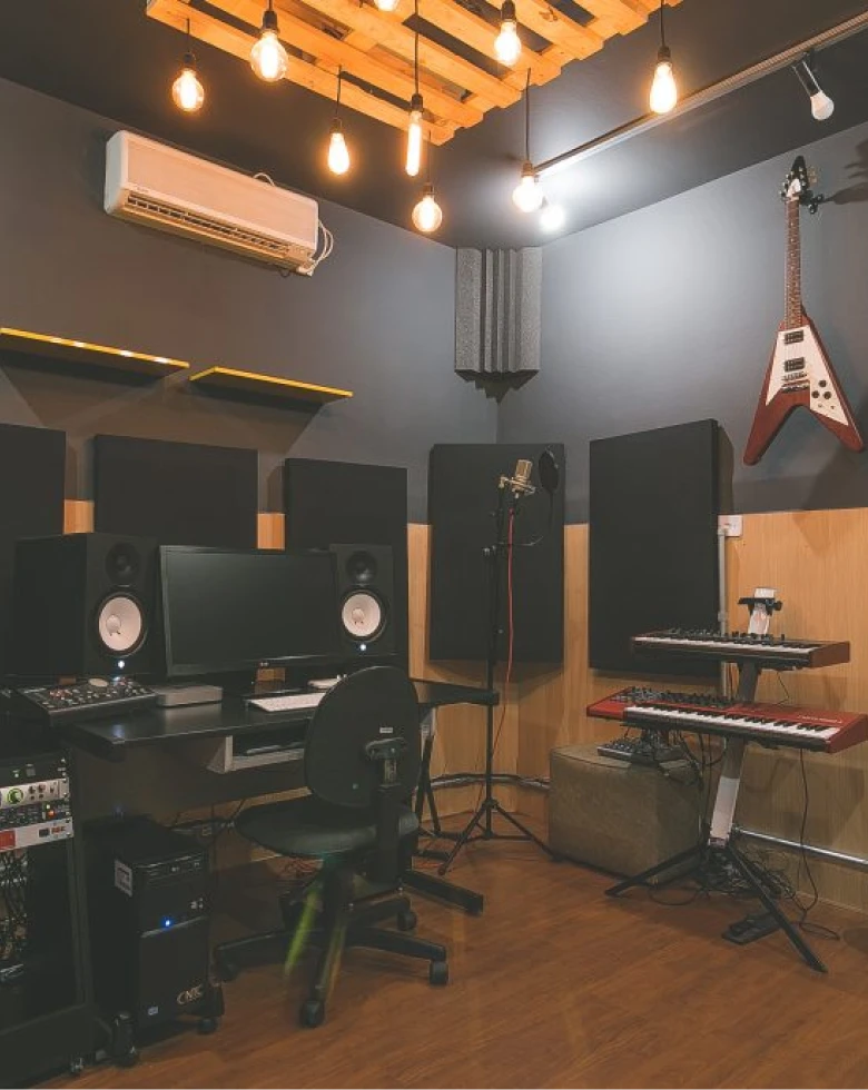 Interior de estúdio de gravação com paredes acústicas, microfone, teclado, guitarra vermelha na parede e cadeira de escritório. Decoração inclui iluminação quente e paletes no teto.
