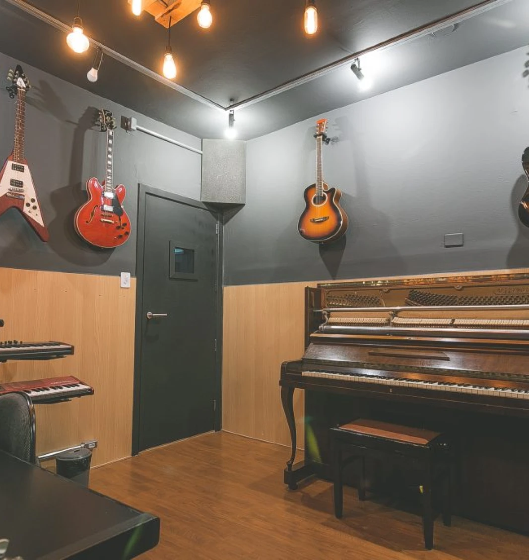 Interior de estúdio de gravação com paredes acústicas, microfone, teclado, guitarra vermelha na parede e cadeira de escritório. Decoração inclui iluminação quente e paletes no teto.